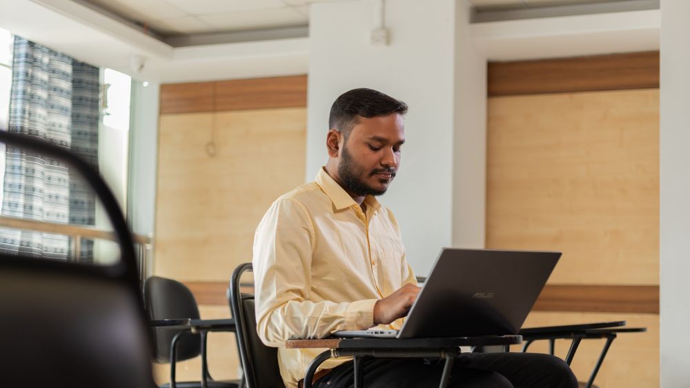 A student sitting at a desk using a laptop in a bright classroom