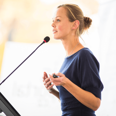 A woman standing at a microphone, making a presentation. She is holding a clicker device.