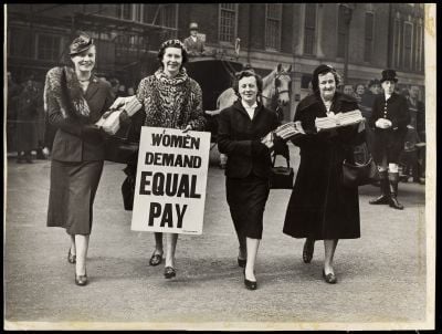 Four women hold up a sign stating 'Women Demand Equal Pay'