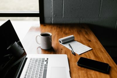 Laptop on a desk with a mug of coffee, a phone and a pad of paper. 