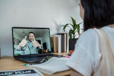 Female student looking at a laptop with a smiling man on the screen looking back at her, with his thumbs up 